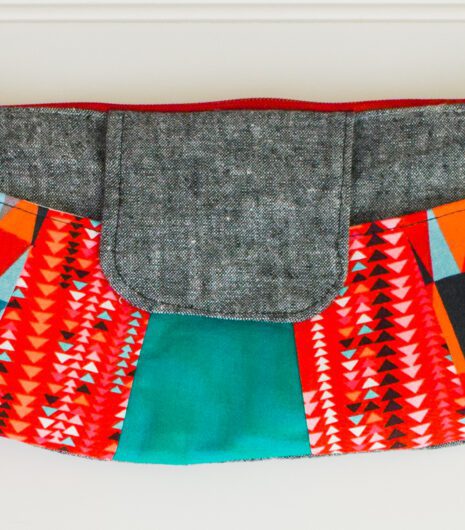 A colorful clutch purse with a gray flap, red zipper, and vibrant geometric patterns in red, orange, turquoise, black, and white on the sides, displayed against a white background.