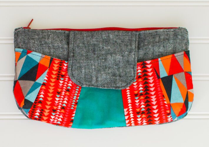 A colorful clutch purse with a gray flap, red zipper, and vibrant geometric patterns in red, orange, turquoise, black, and white on the sides, displayed against a white background.