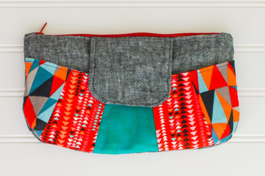 A colorful clutch purse with a gray flap, red zipper, and vibrant geometric patterns in red, orange, turquoise, black, and white on the sides, displayed against a white background.