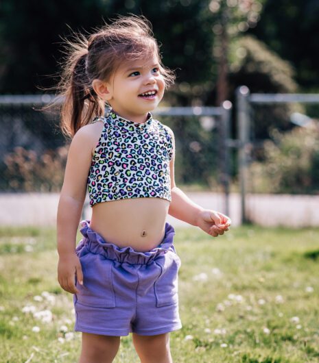 A toddler wearing a patterned crop top and purple shorts is grinning. She is standing in a grassy yard.