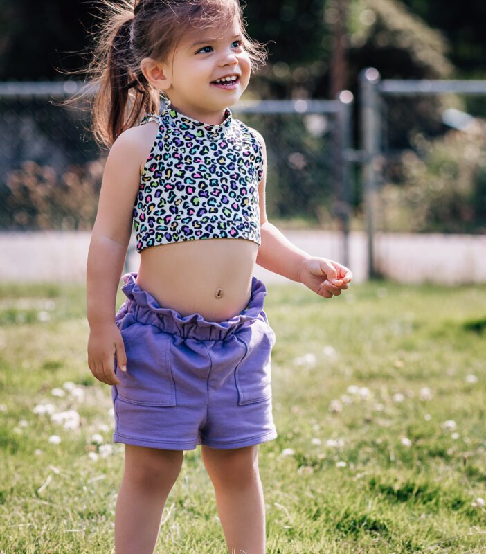A toddler wearing a patterned crop top and purple shorts is grinning. She is standing in a grassy yard.