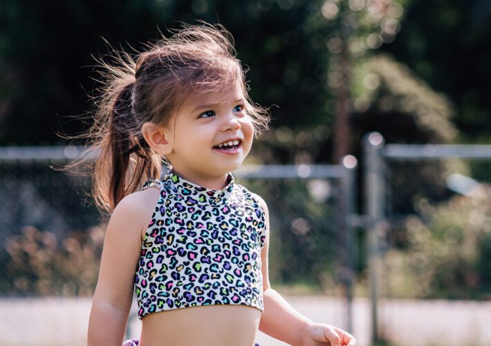 A toddler wearing a patterned crop top and purple shorts is grinning. She is standing in a grassy yard.