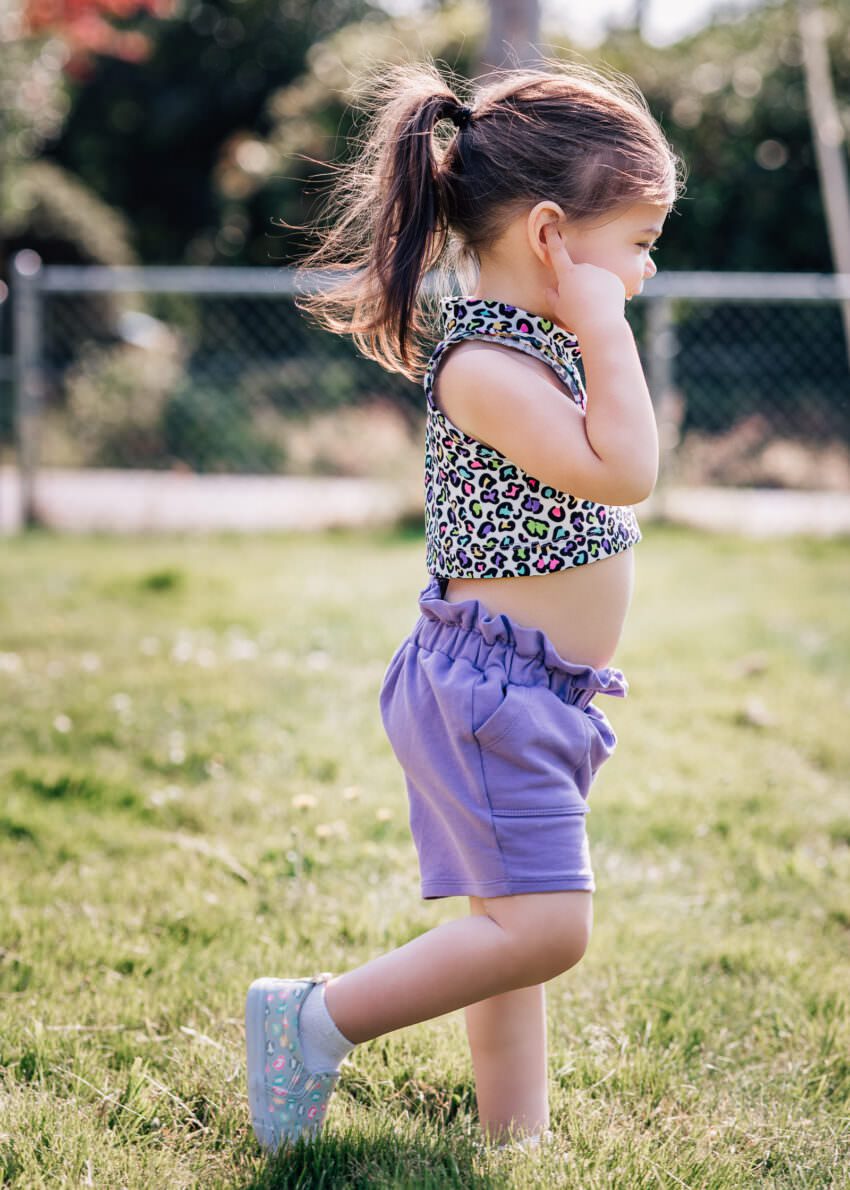 A toddler wearing a patterned crop top and purple shorts is walking through a grassy yard.