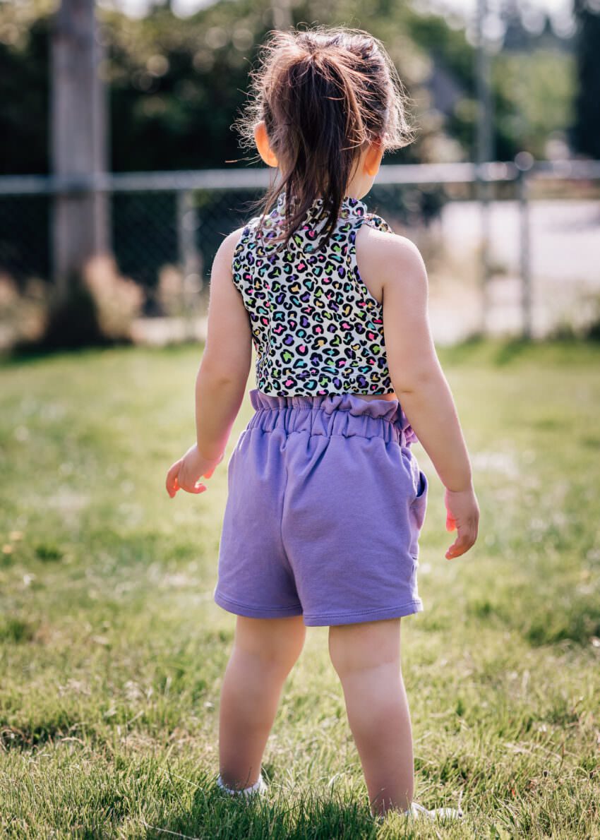 A toddler wearing a patterned crop top and purple shorts is posing to show off her outfit.