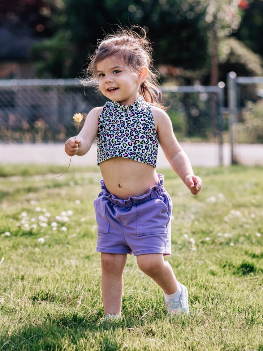 A toddler wearing a patterned crop top and purple shorts smiles while holding a yellow flower. She is standing in a grassy yard.